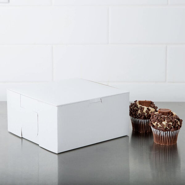A plain white bakery box next to two chocolate chip cupcakes on a stainless steel surface.