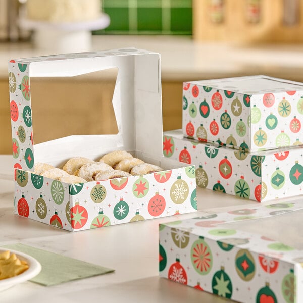 A festive bakery box with a holiday ornament pattern and a clear window on top, partially open to reveal powdered cookies inside, with similar closed boxes stacked nearby.