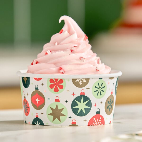 A swirl of pink soft-serve ice cream in a paper cup decorated with colorful holiday ornament designs.