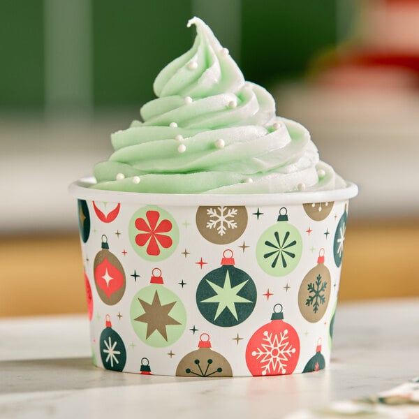 A swirl of light green soft serve topped with white sprinkles in a festive paper cup decorated with colorful ornament designs.