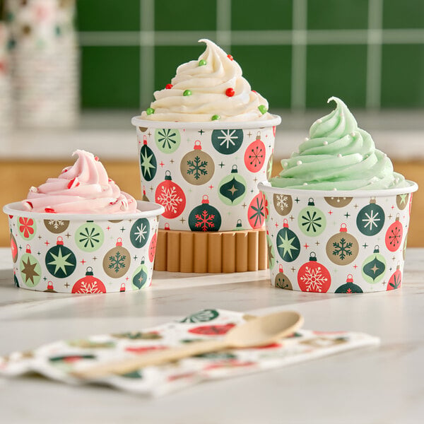 Three paper cups with festive ornament patterns are filled with swirled soft-serve ice cream in pink, white, and green colors, and are placed on a countertop with a napkin and wooden spoon in the foreground.