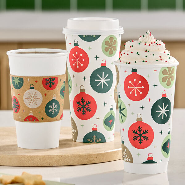 Three festive paper cups with holiday ornament designs, one topped with whipped cream and sprinkles, are displayed on a counter.