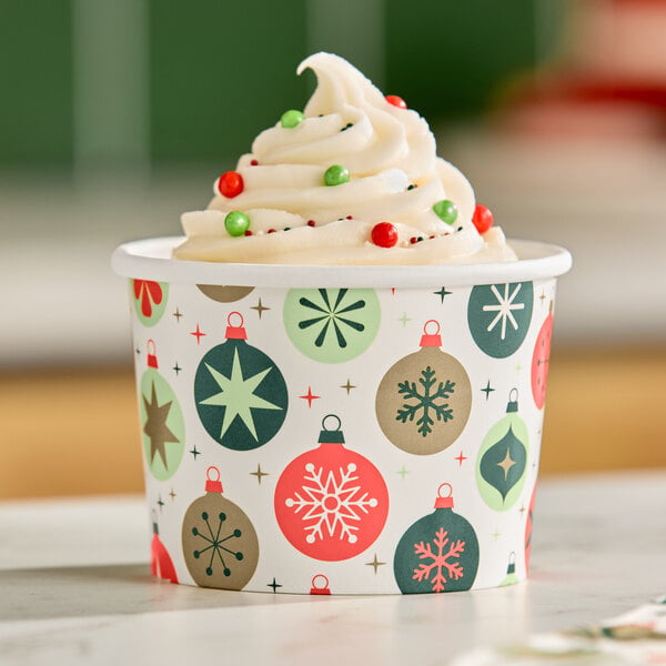 A swirl of vanilla soft serve topped with red and green sprinkles in a festive paper cup decorated with Christmas ornament designs.