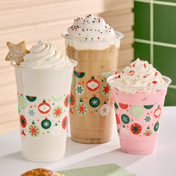 Three festive cold beverages in clear plastic cups decorated with colorful holiday ornament designs, topped with whipped cream and sprinkles, with one featuring a star-shaped cookie.