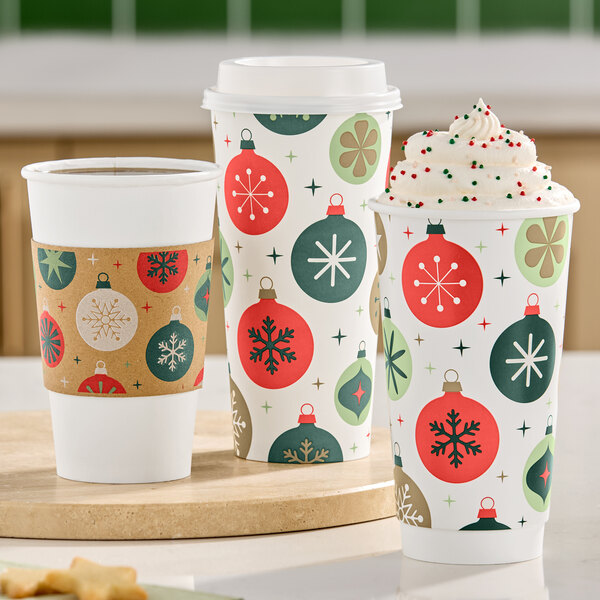 Three festive holiday-themed paper cups are shown, two with lids and one topped with whipped cream and sprinkles, all decorated with colorful ornament patterns.