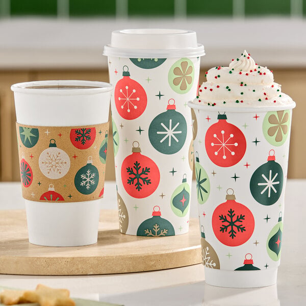 Three festive holiday-themed paper cups are shown, two with lids and one topped with whipped cream and sprinkles, all decorated with colorful ornament patterns.