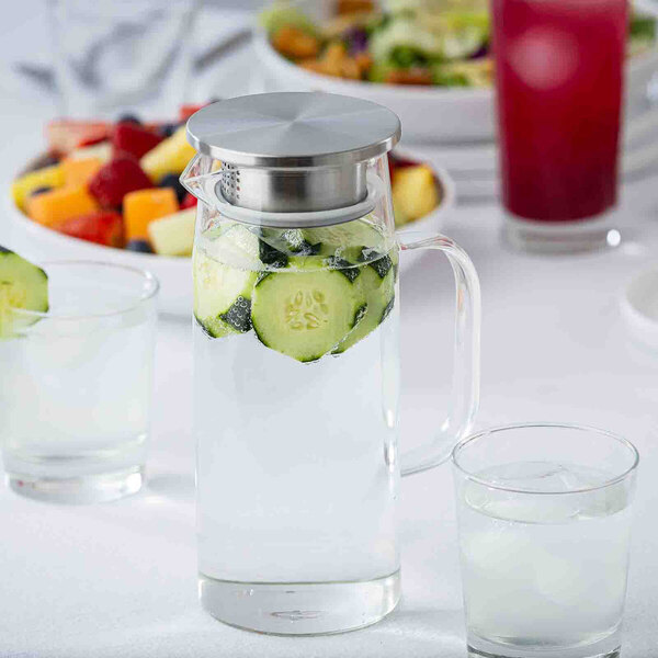 A clear glass pitcher with a stainless steel lid, filled with water and cucumber slices.