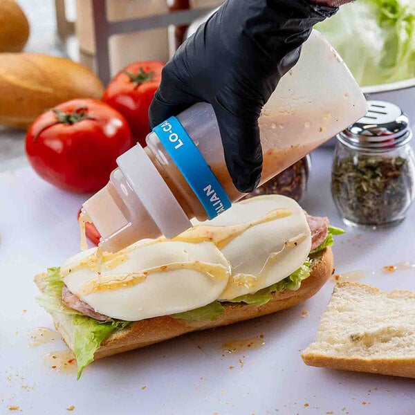 A clear plastic squeeze bottle with a wide mouth and dual-tip top being used to dispense sauce onto a sandwich.