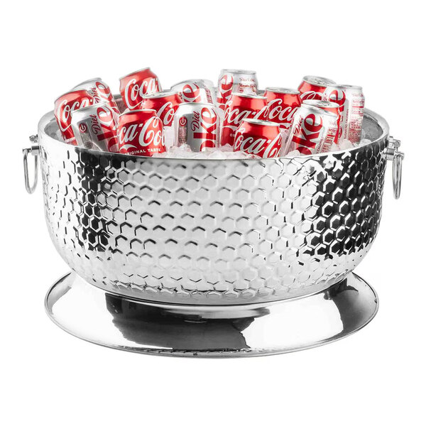 A large round stainless steel beverage tub with a honeycomb pattern, filled with cans of soda and set on a matching base.