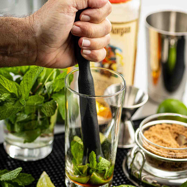A black ABS plastic muddler being used to crush mint and lime in a glass.