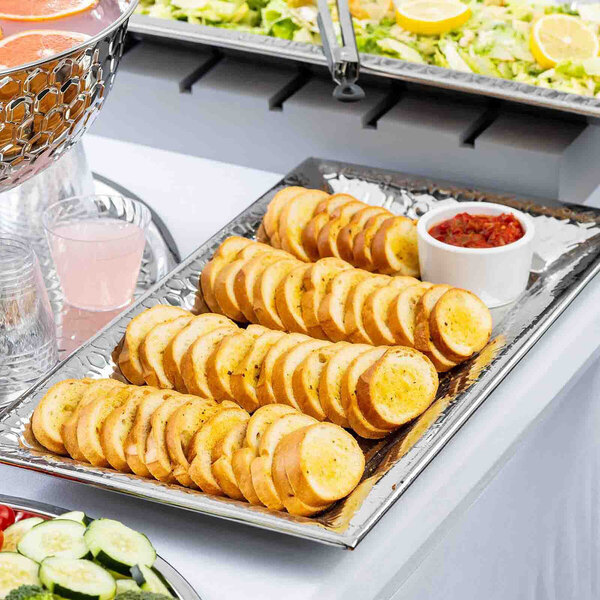 A rectangular stainless steel serving tray with a honeycomb pattern, holding sliced bread and a small bowl of dip.