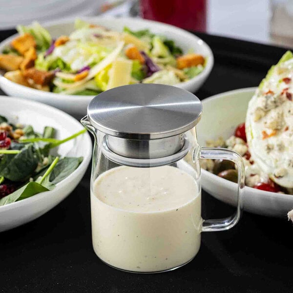 A glass pitcher with a stainless steel lid, filled with a creamy dressing, surrounded by bowls of salad.