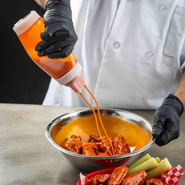 A dual-tip squeeze bottle being used to dispense sauce onto chicken wings in a metal bowl.