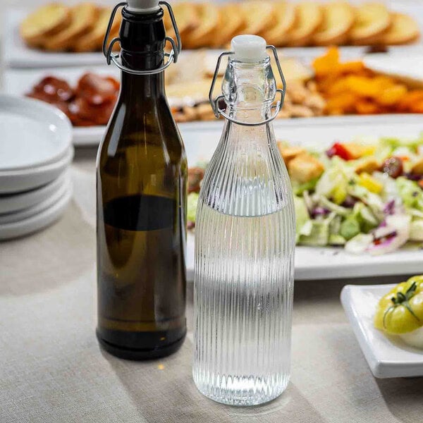 A clear fluted glass carafe with a swing top lid, shown on a table next to another bottle and plates of food.