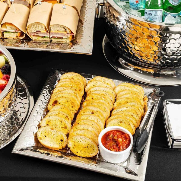 A square stainless steel serving tray with a honeycomb pattern, holding slices of toasted bread and a small bowl of salsa.
