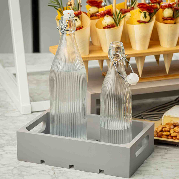 A gray wood serving and display crate holding two ribbed glass bottles with swing-top lids.