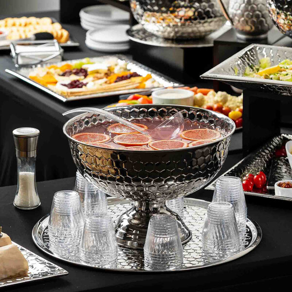A large round stainless steel punch bowl with a honeycomb pattern, filled with punch and garnished with citrus slices, surrounded by clear plastic cups on a serving tray.