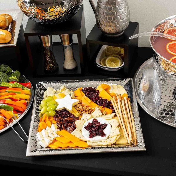 A square stainless steel serving tray with a honeycomb pattern, filled with assorted cheeses, crackers, nuts, dried fruits, and breadsticks.