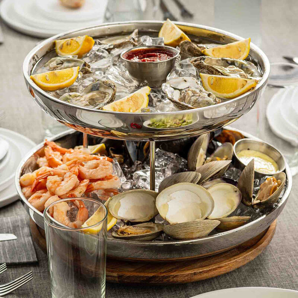 A two-tier stainless steel seafood tower filled with ice, oysters, clams, shrimp, lemon wedges, and dipping sauces.