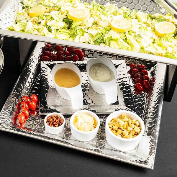A rectangular stainless steel serving tray with a honeycomb pattern, holding various salad ingredients and dressings.