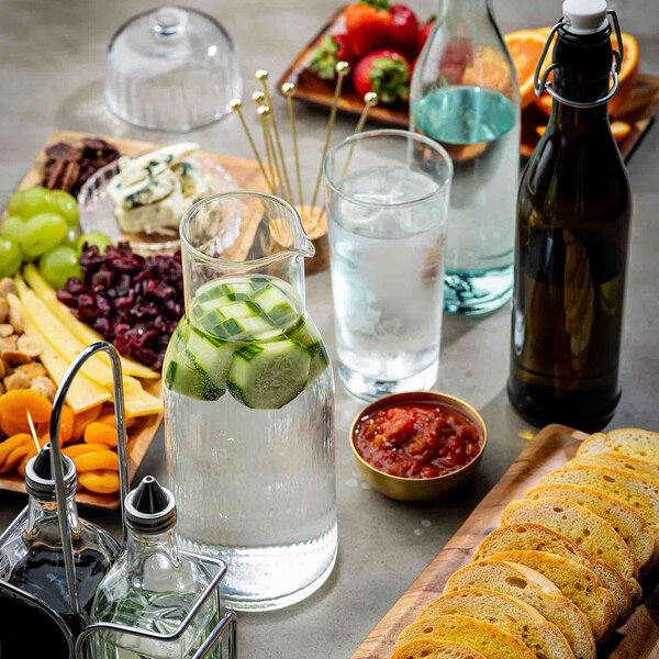 A 34 oz. pebbled glass carafe filled with water and cucumber slices, placed on a table with assorted snacks and drinks.