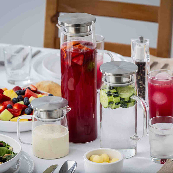 A glass pitcher with a stainless steel lid, shown filled with a red beverage and fruit slices on a dining table.