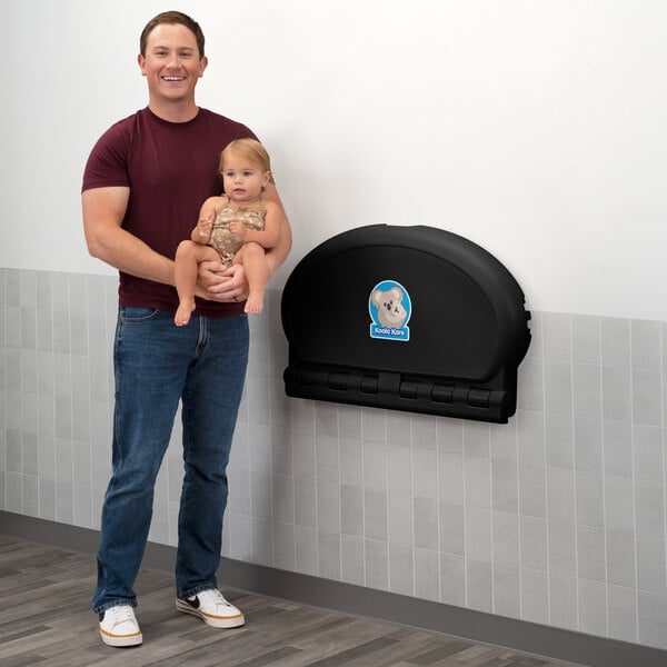 A man holding a baby stands next to a black, wall-mounted, horizontal oval baby changing station attached to a tiled wall.