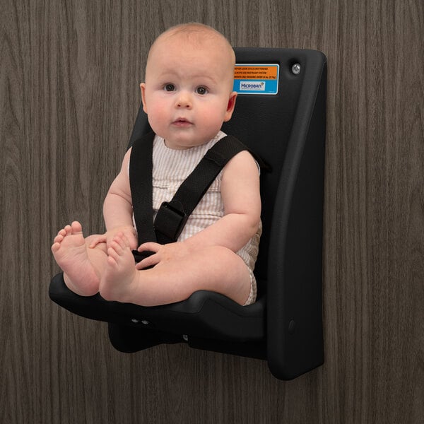 A baby is seated and secured with straps in a black wall-mounted child safety seat against a wood-patterned wall.