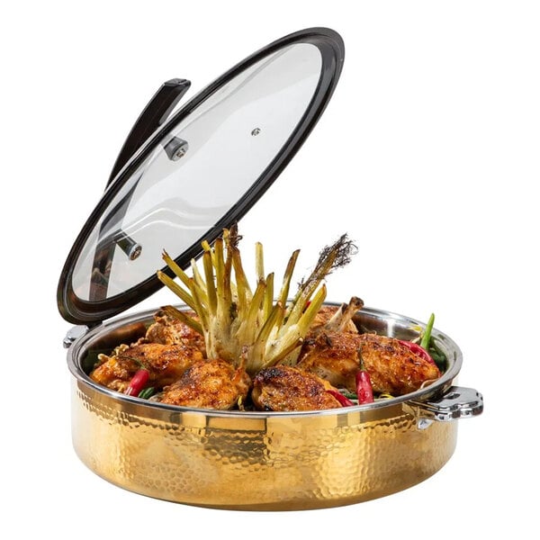 A round, gold-colored hammered steel buffet server with a glass lid, containing cooked chicken pieces and garnished with vegetables.
