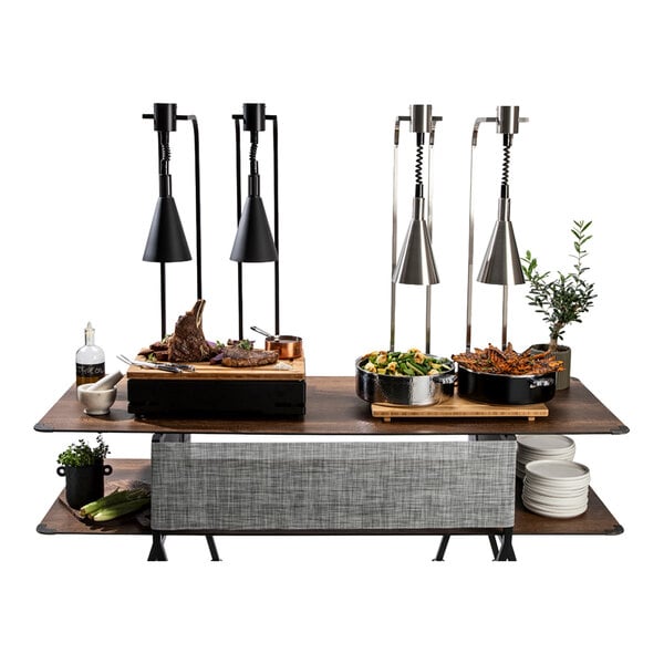 A buffet table setup with four freestanding heat lamps positioned over various dishes, including meat, vegetables, and garnishes, with plates and decorative plants on the side.