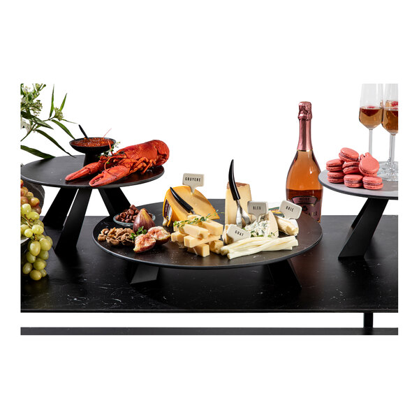 A round, gray riser with a modern black angled leg, used to elevate food displays such as cheese, lobster, and desserts.