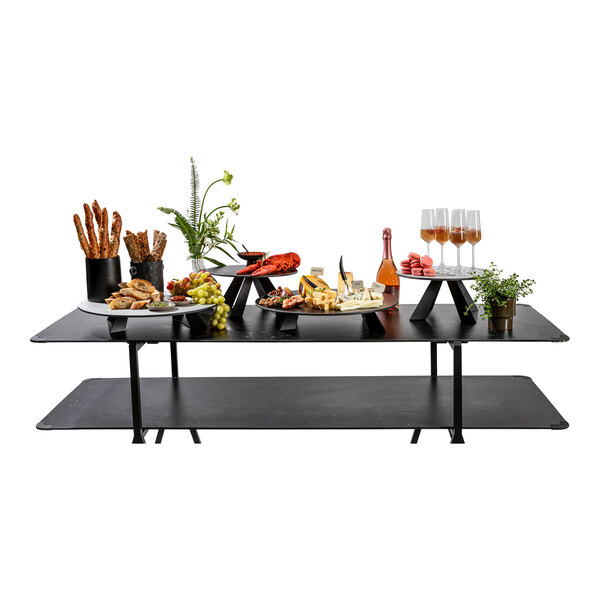 A black rectangular table with various food items, drinks, and decorative plants arranged on top, including elevated round risers displaying cheese, bread, fruit, and glasses.