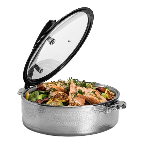 A round hammered steel buffet server with a glass lid, containing cooked salmon, vegetables, and lemon slices.