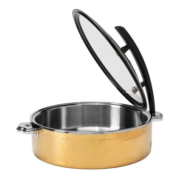 A round, gold-colored hammered steel buffet server with a glass lid that is propped open, featuring two side handles.