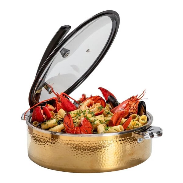 A round, gold hammered steel buffet server with a glass lid, filled with pasta, lobster, and mussels.