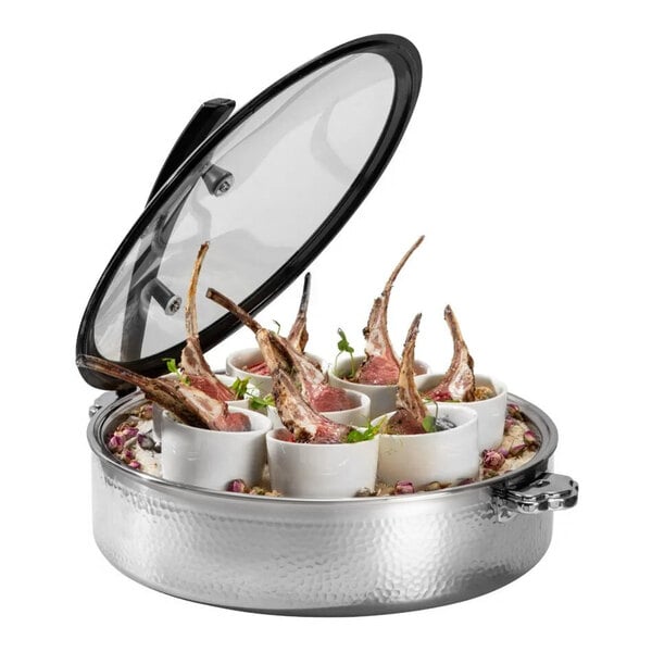 A round, hammered steel buffet server with a glass lid, containing several white cups each holding a piece of cooked meat with bones, garnished with herbs.