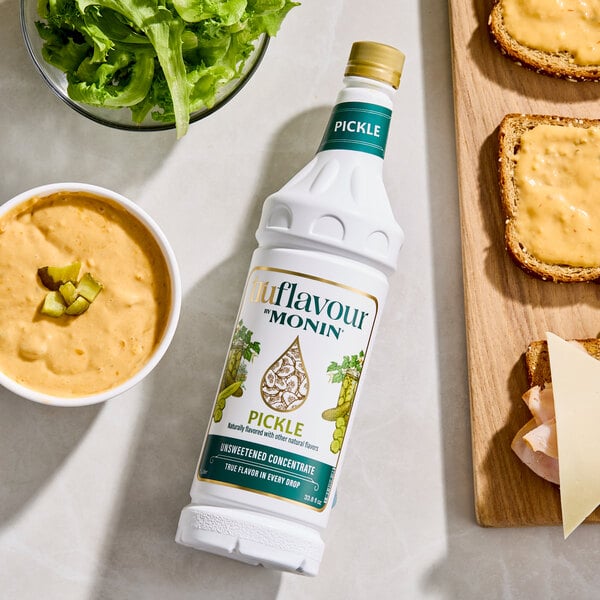 A bottle labeled 'Pickle' flavor concentrate is placed on a countertop surrounded by a bowl of leafy greens, a bowl of creamy dip with pickle pieces, and slices of bread with cheese on a wooden board.