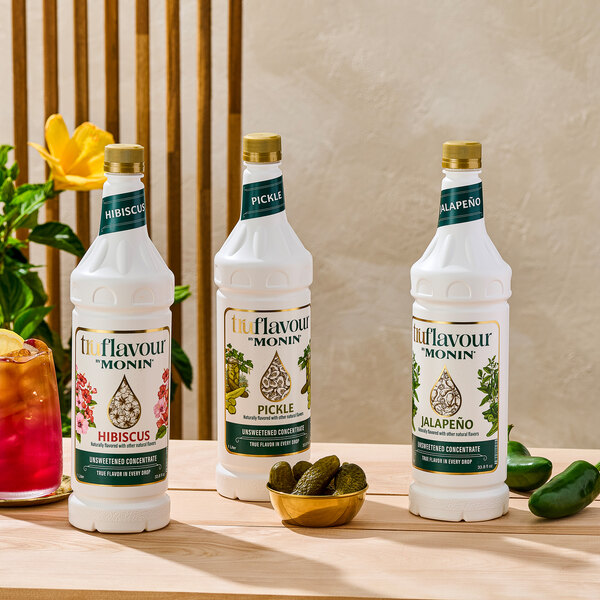 Three white bottles of Monin TruFlavour unsweetened concentrate in hibiscus, pickle, and jalapeño flavors are displayed on a wooden surface, with a bowl of pickles, a glass of red beverage, and some jalapeños nearby.