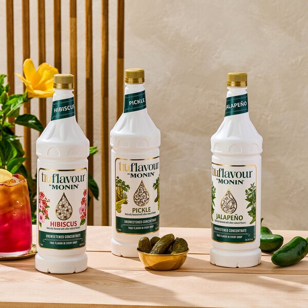 Three white bottles of TruFlavour by Monin unsweetened culinary flavor concentrates (Hibiscus, Pickle, and Jalapeño) are displayed on a wooden surface, with a bowl of pickles, a glass of red beverage, and some jalapeños nearby.