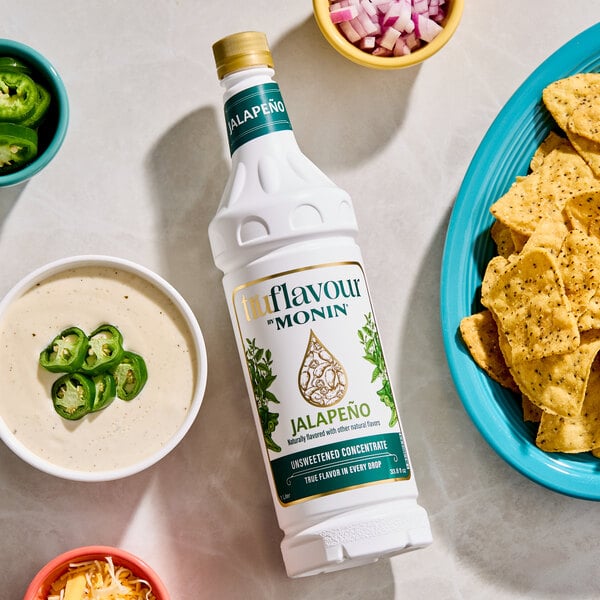 A bottle of Monin Jalapeño unsweetened concentrate is placed on a table surrounded by a bowl of tortilla chips, a bowl of cheese sauce with sliced jalapeños, and small bowls of chopped onions, shredded cheese, and sliced jalapeños.