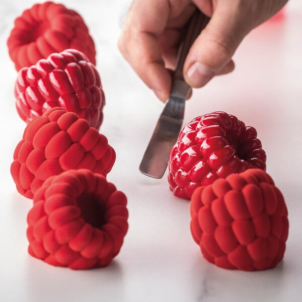A hand is using a spatula to handle realistic raspberry-shaped desserts, some with a shiny finish and others with a matte texture, arranged on a white surface.