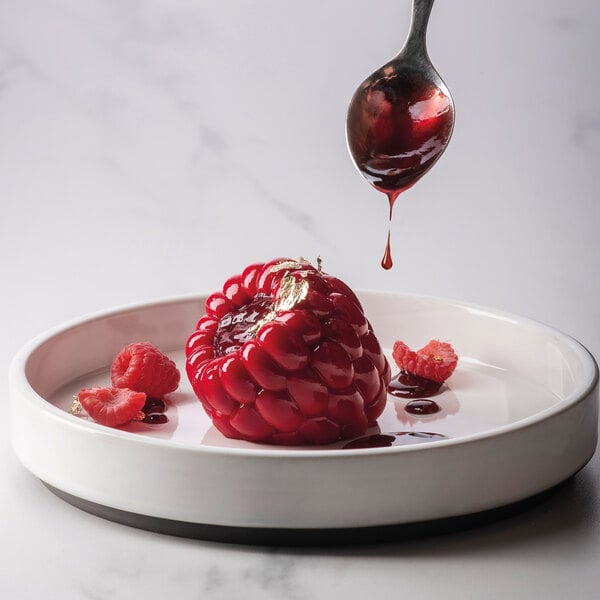 A dessert shaped like a raspberry with a glossy red finish, garnished with gold leaf and fresh raspberries, is being drizzled with red sauce from a spoon on a white plate.