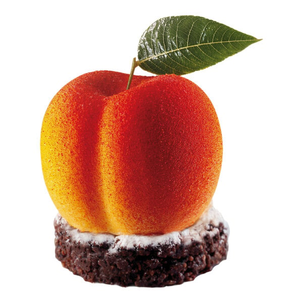 A dessert shaped and colored like a peach with a green leaf on top, sitting on a round chocolate base dusted with powdered sugar.