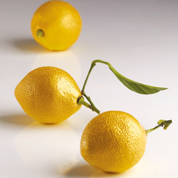 Three realistic lemon-shaped objects with textured yellow surfaces, one with a green leaf attached, are displayed on a smooth white surface.