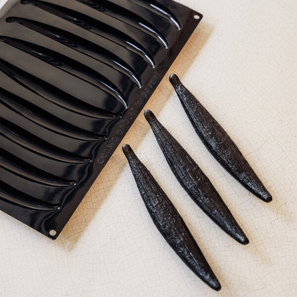 A black silicone mold with elongated, curved cavities is shown next to three dark, glossy, bean-shaped molded items.