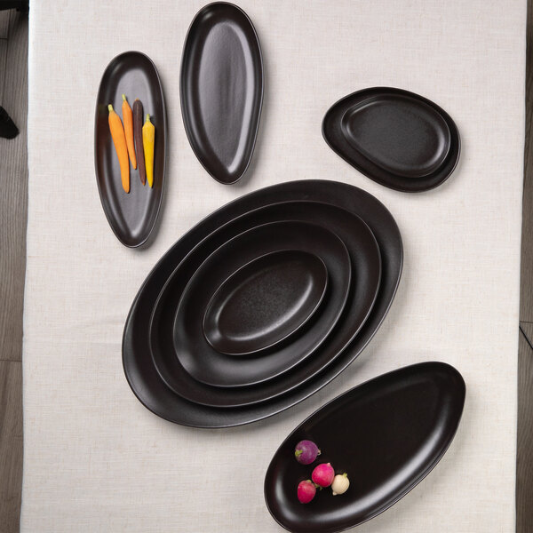 Several matte black, oval and organic-shaped stoneware plates are arranged on a light-colored tablecloth, with a few vegetables placed on two of the plates.