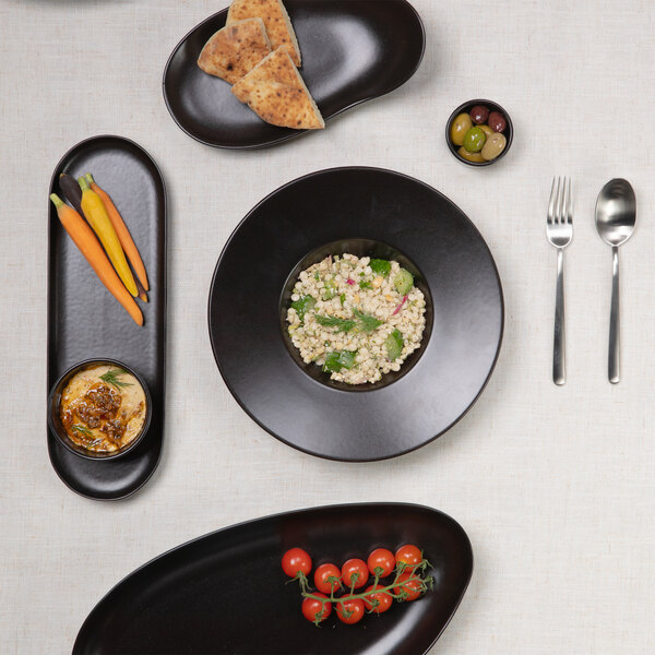 A top-down view of a table setting with black stoneware dishes containing various foods, including carrots, cherry tomatoes, flatbread, olives, a grain salad, and a dip, arranged on a light tablecloth with a fork and spoon.
