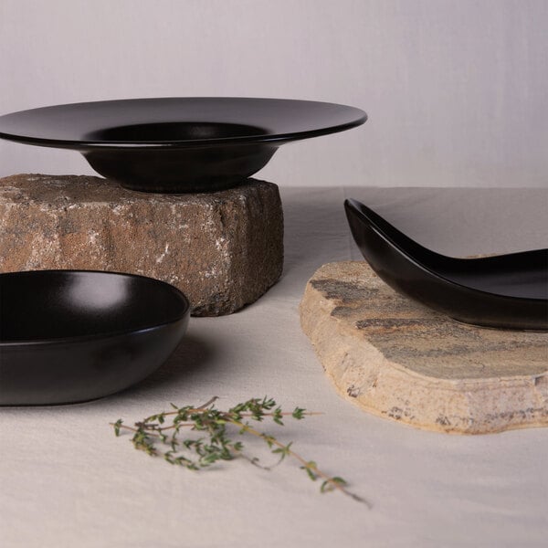 Three matte black ceramic bowls of varying shapes are displayed on stone slabs with a sprig of herbs nearby.