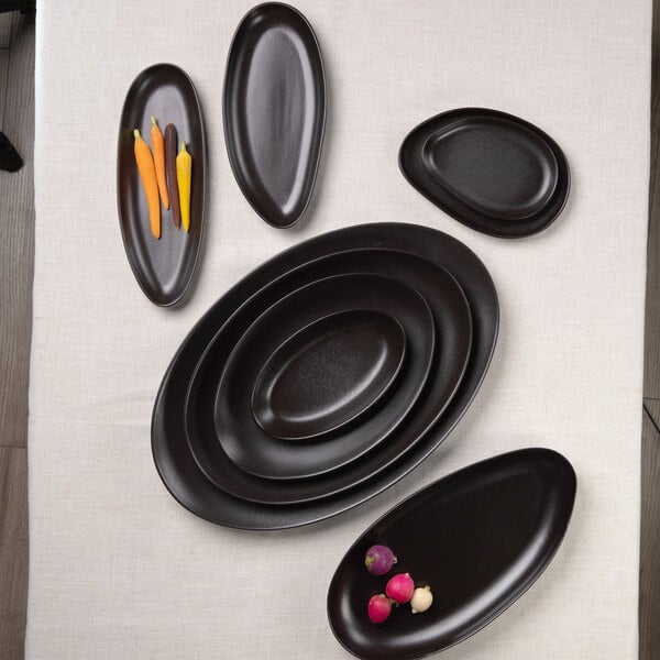 A set of matte black, oval and organically shaped stoneware bowls and platters arranged on a light-colored tablecloth, with a few vegetables placed on two of the dishes.