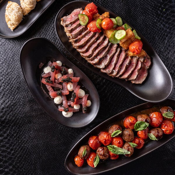 A top-down view of several black oval trays with assorted gourmet foods, including sliced steak with vegetables, a dish with diced ingredients and sauce, and another with roasted tomatoes and herbs, all arranged on a textured black surface.